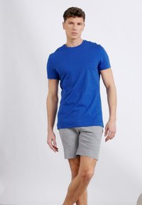 Άνδρας που στέκεται φορώντας μπλε κοντομάνικη μπλούζα και γκρι σορτς σε απλό ανοιχτό φόντο.