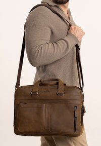 Bruine leren aktetas met twee handvatten aan de bovenkant en een afneembare schouderriem, voorzien van meerdere rits- en insteekvakjes voor organisatie.