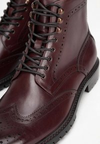 Burgundské kožené boty s brogue detaily, černými šňůrkami, bronzovými očky a odolnou gumovou podrážkou, zdůrazňující klasické řemeslné zpracování.