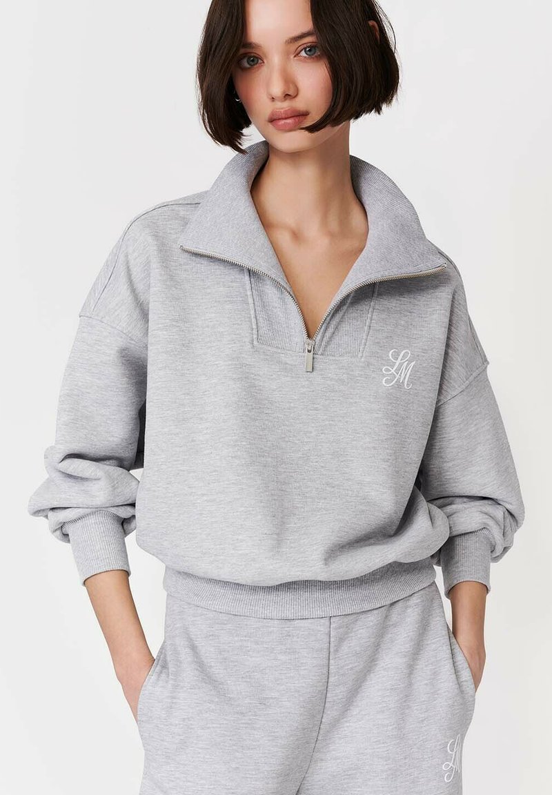 Tânără purtând un hanorac gri deschis cu fermoar și pantaloni de trening asortați, cu inițiale albe brodate, cu mâinile în buzunare, pe un fundal neutru.