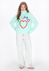 Mintgrüner Kabelstrickpullover mit bunten gestickten Buchstaben und Herzdesign, kombiniert mit hellblauen, ausgestellten Jeans und rosa Schuhen.