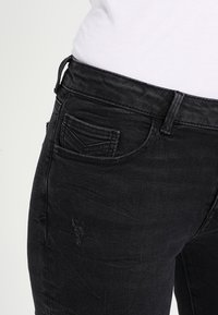 Czarne dżinsy o zniszczonym wyglądzie, z małą kieszenią z przodu i widocznymi detalami szwów.