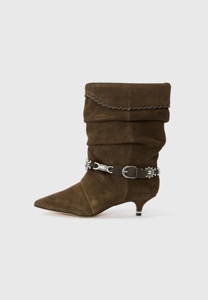 Stivaletti a caviglia in suede marrone con design morbido, punta affusolata e una cintura decorativa in argento attorno alla caviglia. Presentano un tacco basso a forma di kitten.