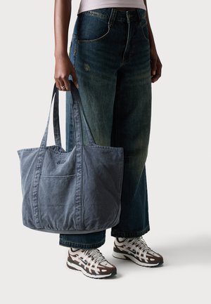 Personne portant un jean large et des baskets, tenant un grand sac fourre-tout bleu délavé avec double poignées.