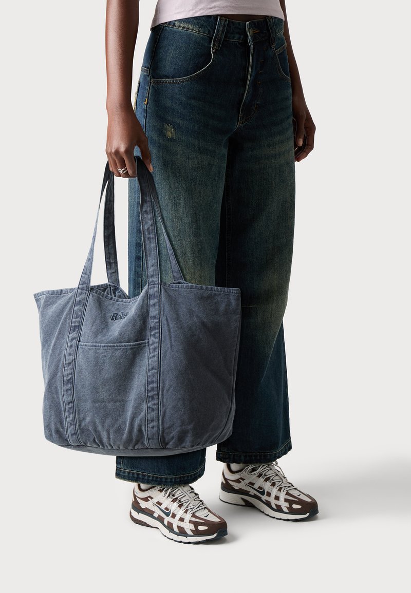 En person som har på seg vidde jeans og sneakers, holder en stor falmet blå totebag med doble håndtak.