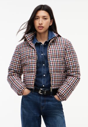 Vrouw draagt een korte donsjacket met een meerkleurig ruitpatroon over een denim blouse en blauwe jeans, handen in de zakken, gezicht naar voren.