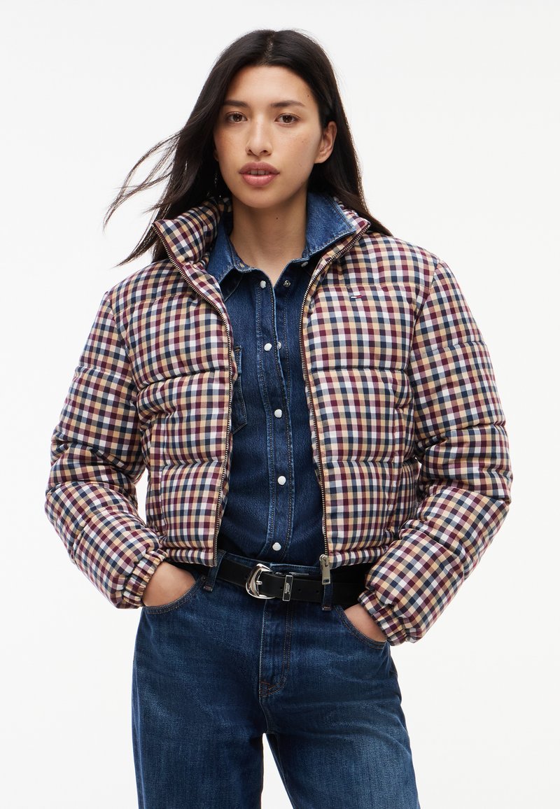 Femme portant une doudoune courte à motif carreaux multicolores par-dessus une chemise en jean et un jean bleu, mains dans les poches, face à l'avant.