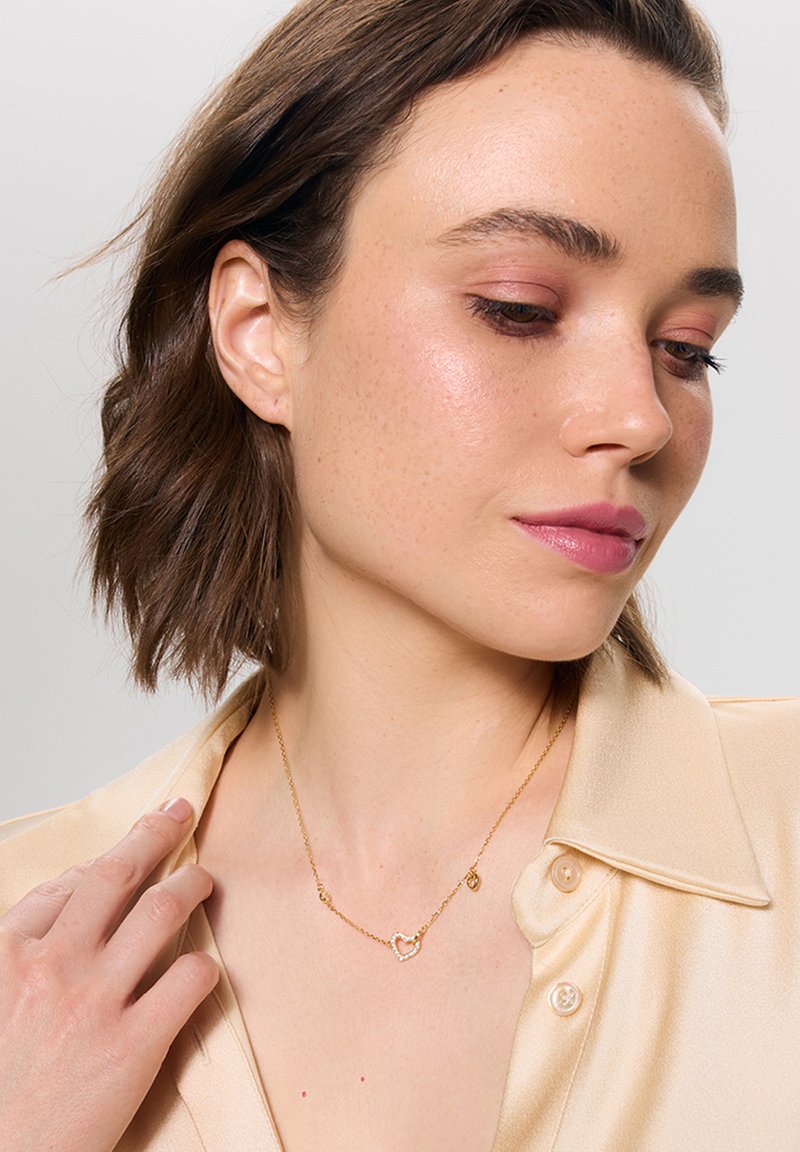 Złoty naszyjnik z wisiorkiem w kształcie serca i małym charms'em. Noszony z jedwabną, beżową koszulą z kołnierzykiem, o gładkiej fakturze i delikatnym połysku.