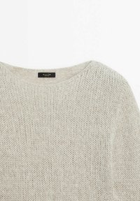 Pull en tricot beige clair avec un col rond et un motif fin texturé, montrant une étiquette avec le nom de la marque à l'intérieur du col.