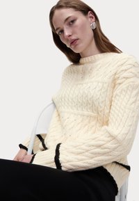 Maglione crema a trecce con collo e orlo a coste, caratterizzato da bordi neri a contrasto su polsini e fondo. Tessuto liscio e strutturato.