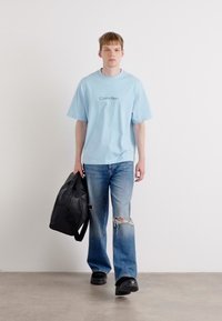 Lyseblå oversized t-skjorte med "Calvin Klein"-logo, kombinert med slitte blå jeans og svarte slip-on sko, mens personen holder en svart veske.