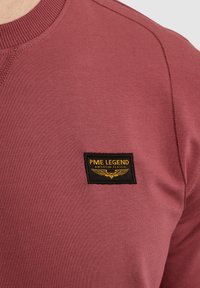 Maroon dukserica s rebrastim izrezom, teksturiranom tkaninom i istaknutim crnim logom s zlatnim natpisom i grafikom krila.
