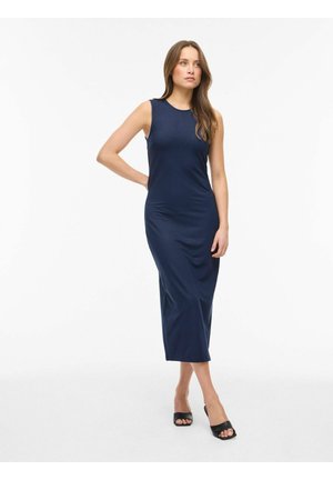 Kobieta w bezrękawnikowej, granatowej sukience midi oraz czarnych, otwartych na palce szpilkach stoi z jedną ręką na biodrze na białym tle.