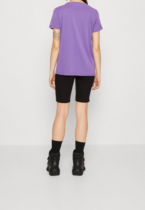 Osoba ubrana w fioletową koszulkę z krótkim rękawem, czarne spodenki, czarne skarpetki i czarne buty, pokazująca tatuaż pajęczyny na lewym ramieniu.