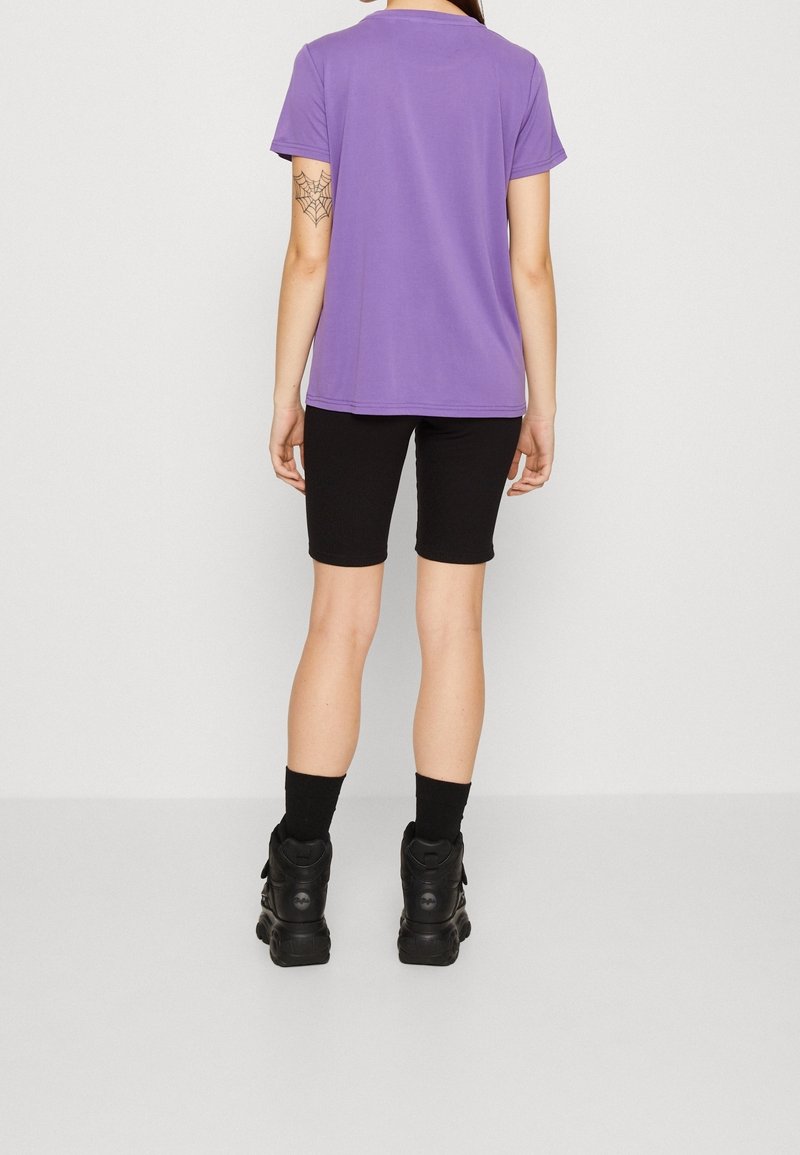 Personne portant un t-shirt violet à manches courtes, un short noir, des chaussettes noires et des bottes noires, montrant un tatouage en forme de toile d'araignée sur son bras gauche.