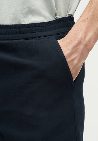 Χέρι που έχει μπει στην δεξιά τσέπη μαύρων παντελονιών με ελαστική μέση, φορεμένων με ανοιχτό γκρι πουκάμισο.