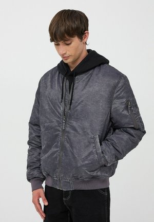 Giacca bomber grigia con finitura opaca, polsini a coste, due tasche laterali e una tasca utility con zip sul braccio. Indossata sopra un felpa nera.