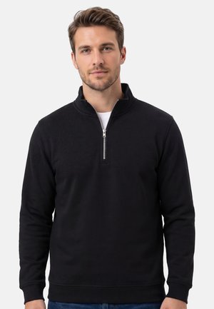 Man draagt een zwarte pullover met kwart ritssluiting en een blauwe spijkerbroek, staat tegen een effen lichtgrijze achtergrond, kijkt recht vooruit.