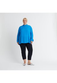 Blaues Langarmoberteil mit hochgeschlossenem Kragen, kombiniert mit schwarzen, knöchellangen Hosen. Die Schuhe sind silber mit glänzender Textur.