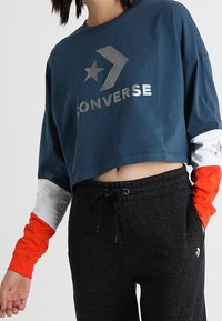 Krátká modrá mikina se stříbrným logem "Converse", hvězdou a designem chevron, s dlouhými rukávy v oranžové a bílé barvě, doplněná černými tepláky.