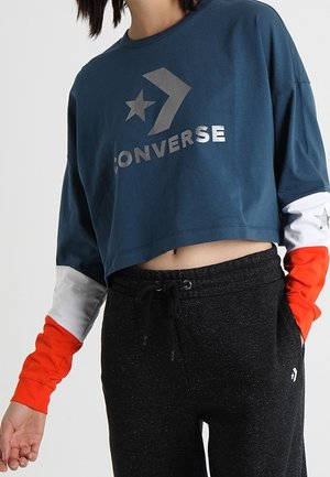 Gekürzter blauer Sweatshirt mit silbernem "Converse"-Logo, Stern und Chevron-Design, ausgestattet mit langen Ärmeln in Orange und Weiß, kombiniert mit schwarzen Jogginghosen.