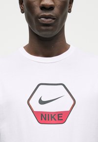 Κοντινό πλάνο ενός ατόμου που φοράει λευκό πουκάμισο με μαύρο και κόκκινο εξαγωνικό λογότυπο της Nike στο στήθος.