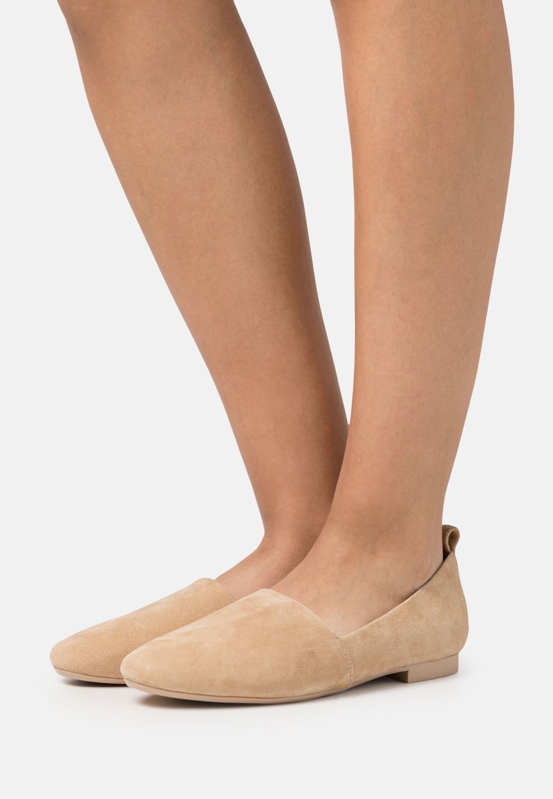 Pieds chaussés de ballerines pointues en daim beige à petit talon, photographiés sur un fond clair uni.