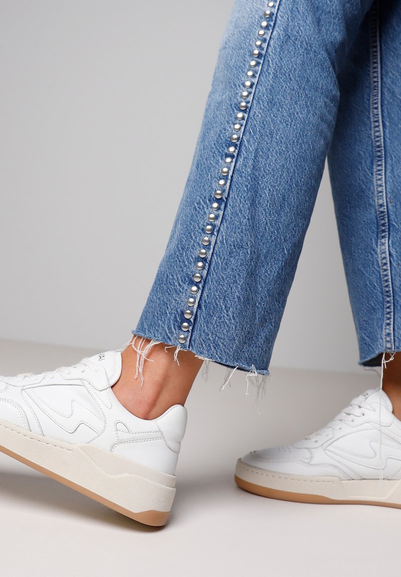 Weiße Sneakers mit strukturierten Oberflächen, kombiniert mit blauen Jeans mit Fransen und silbernen Nieten entlang der Seitennaht.