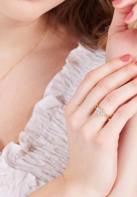 Gouden ring met een centrale heldere steen en twee kleine stenen aan elke kant, vastgehouden tussen vingers met lichtroze nagellak. Rimpelig wit stoffen achtergrond.