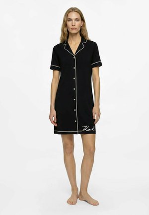 Femme portant une robe noire à manches courtes avec des boutons, un liseré blanc et "Karl" brodé près de l'ourlet, debout pieds nus sur fond blanc.