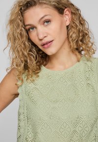Top senza maniche di un verde chiaro realizzato in pizzo con motivi intricati, dotato di bordi scallop e un design texture.