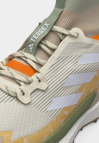 Close-up van een beige en oranje Adidas Terrex trail-schoen met de nadruk op de veters, logo-strepen en het geprofileerde zoolpatroon.
