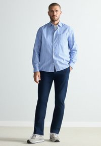 Camasa cu dungi albastre și albe, pantaloni bleumarin și adidași gri. Camasa are un buzunar frontal și mâneci lungi.