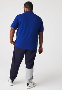Homme portant un polo bleu à manches courtes, un pantalon bicolore bleu marine et gris, et des baskets blanches, debout de dos face à la caméra.