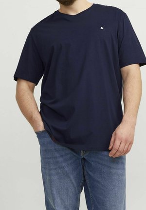 Mężczyzna noszący gładki, ciemnogranatowy T-shirt z małym białym logo ampersanda na piersi oraz niebieskie dżinsy, stojący z jedną ręką w kieszeni.