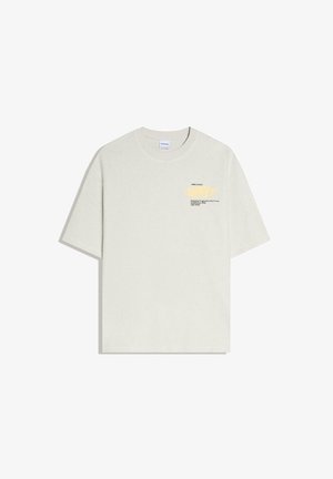 Kremowa koszulka z krótkim rękawem i okrągłym dekoltem, z małym czarnym napisem i jasnżółtym abstrakcyjnym wzorem na lewej części klatki piersiowej.