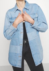 Svetle modrá džínová bunda s knoflíkovým zapínáním, dvěma náprsními kapsami a dlouhými rukávy. Disponuje patchworkovým designem a mírně zaobleným lemem.