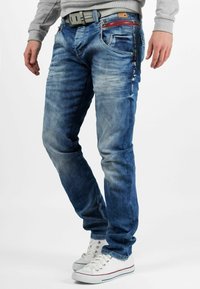 Un jean en denim bleu clair avec des zones décolorées, plusieurs poches, des accents en zip et une fermeture éclair rouge contrastante. Associé à des baskets blanches.