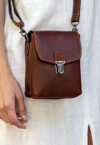 Brown leather crossbody bag with a flap, metal clasp, and adjustable strap. Features visible stitching and a compact rectangular shape.