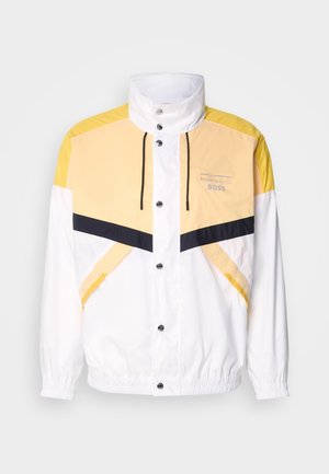 Biely vetrovka s žltými a tmavomodrými akcentmi, s vysokým golierom, šnúrkou na stiahnutie a cvokmi. Ľahký, hladký materiál.