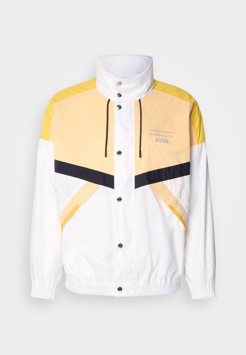Veste coupe-vent blanche avec des accents jaunes et marine, dotée d'un col haut, d'un cordon de serrage et de boutons-pression. Texture légère et lisse.