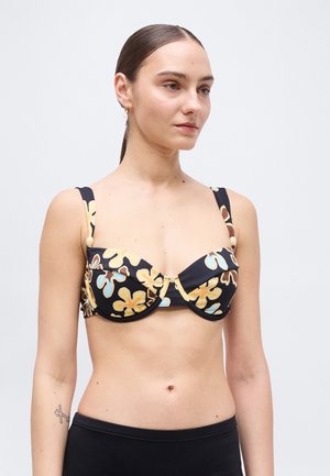 Kobieta w czarnym topie bikini z dużymi żółto-niebieskimi kwiatowymi wzorami, w połączeniu z czarnymi majtkami, na jednolitym białym tle.