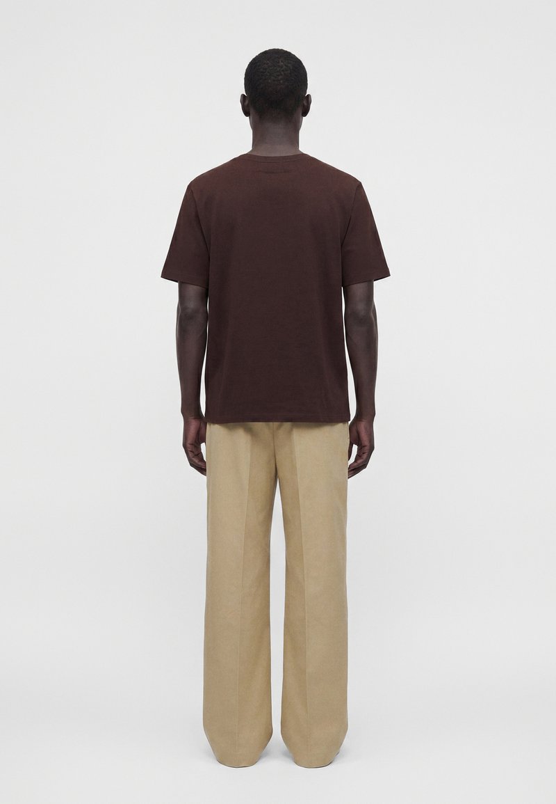Mann, der mit dem Rücken zur Kamera steht, trägt ein dunkelbraunes kurzärmeliges Hemd und beige weite Hosen vor einem schlichten Hintergrund.