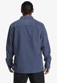 Camisa de corduroy azul con rayas verticales, cuello clásico y puños abotonados. Presenta una zona de hombros en contraste y un diseño de corte relajado.