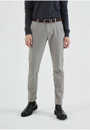 Man draagt grijze slim-fit broek, zwarte riem, donkergroene lange mouw shirt en zwarte leren laarzen, staand tegen een effen achtergrond.