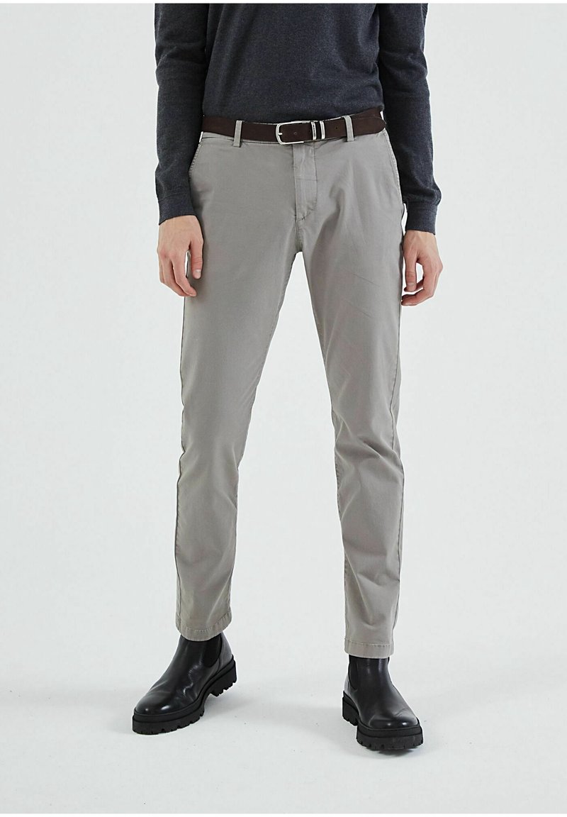 Homme portant un pantalon slim gris, une ceinture noire, une chemise à manches longues gris foncé et des bottes en cuir noires, debout devant un fond uni.