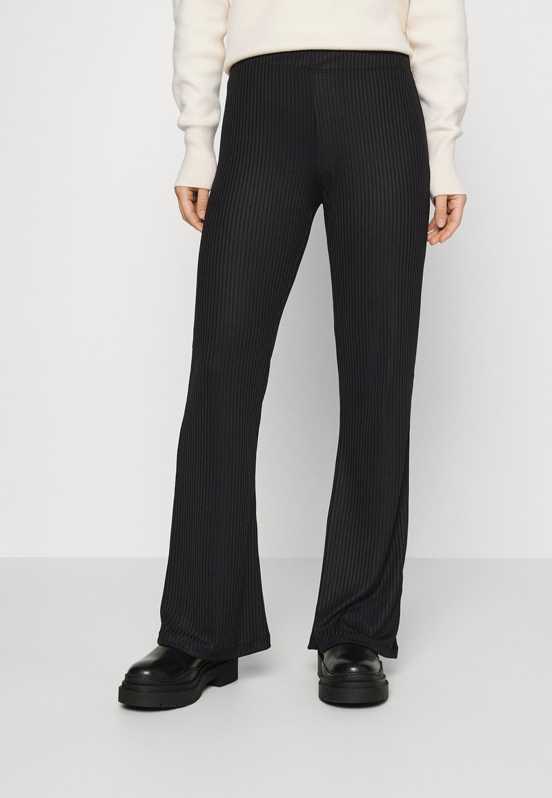 Personne portant un pantalon noir côtelé évasé, un haut à manches longues crème et des chaussures plateformes noires épaisses, debout devant un fond uni.