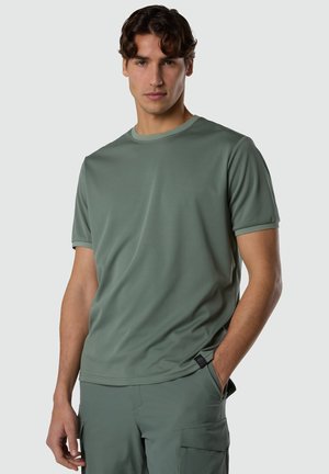 Giovane uomo con capelli castani ondulati che indossa una semplice t-shirt a maniche corte verde salvia e pantaloni cargo abbinati, in piedi con una mano in tasca.