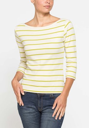 Donna che indossa una camicia bianca con righe orizzontali gialle e maniche a tre quarti, abbinata a jeans blu scuro, mano appoggiata sul fianco.
