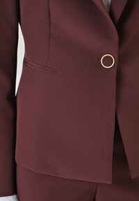Burgunderfarbener Blazer mit tailliertem Design, einem goldenen, runden Knopf und einer glatten Textur. Verfügt über eine einzelne Brusttasche und strukturierte Revers.